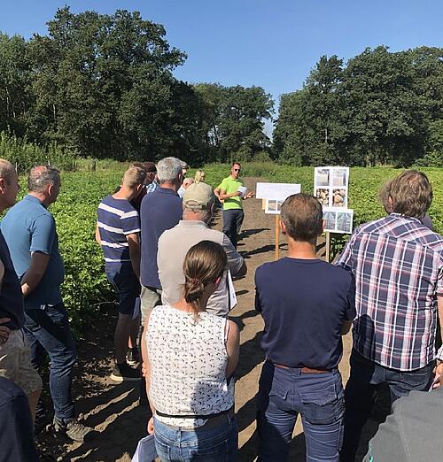 An einem Feldweg steht ein Mann in grünem Poloshirt mit einem Zettel in der Hand. Vor ihm stehen etwa ein Dutzend Zuhörende.